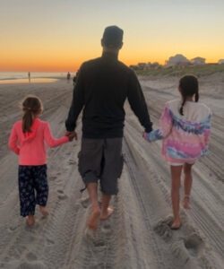 Sean walks on the beach at sunset, holding hands with his two daughters