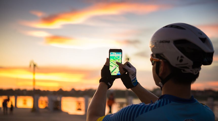 Male cyclist uses hand to hold a smartphone with a running Pokemon Go application at sunset.