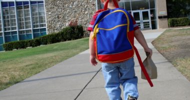 little boy wearing backpack and holding a bag lunch walks toward a school