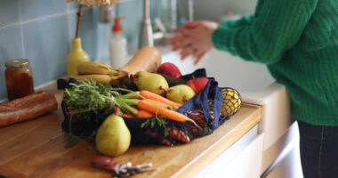 pile of fruits and vegetables on a butcher-block kitchen island, someone washing their hands in the sink in the background