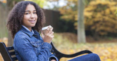 Image of young woman of color enjoying a coffee in a to-go cup on a bench in a park wearing a denim jacket.