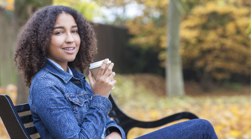 Image of young woman of color enjoying a coffee in a to-go cup on a bench in a park wearing a denim jacket.