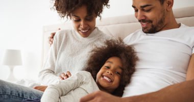 Image of a little girl laughing in bed with her mom and dad.