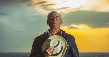 Older man with hat against his chest stands in front of a beautiful golden sunset.
