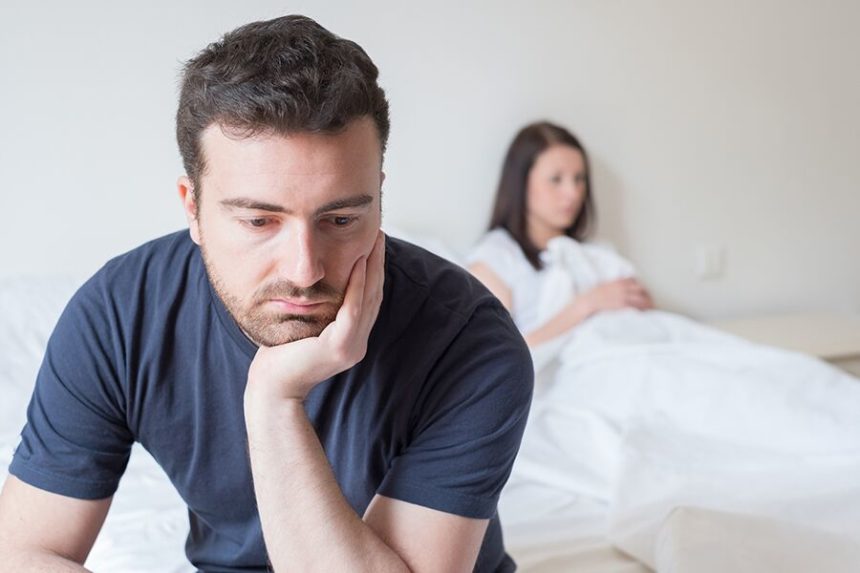 Man sitting on edge of bed looking sad while female partner is in the background