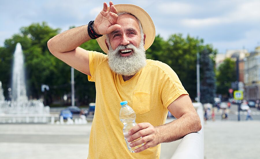 Older man standing in a town square, wiping brow while holding a water bottle