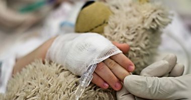 close up of child's hand holding a teddy bear, laying in a hospital bed with an IV insterted