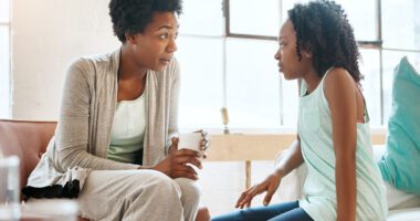 An adult woman talks with a young girl in a living room, showing attention and empathy as they share a serious but caring conversation.