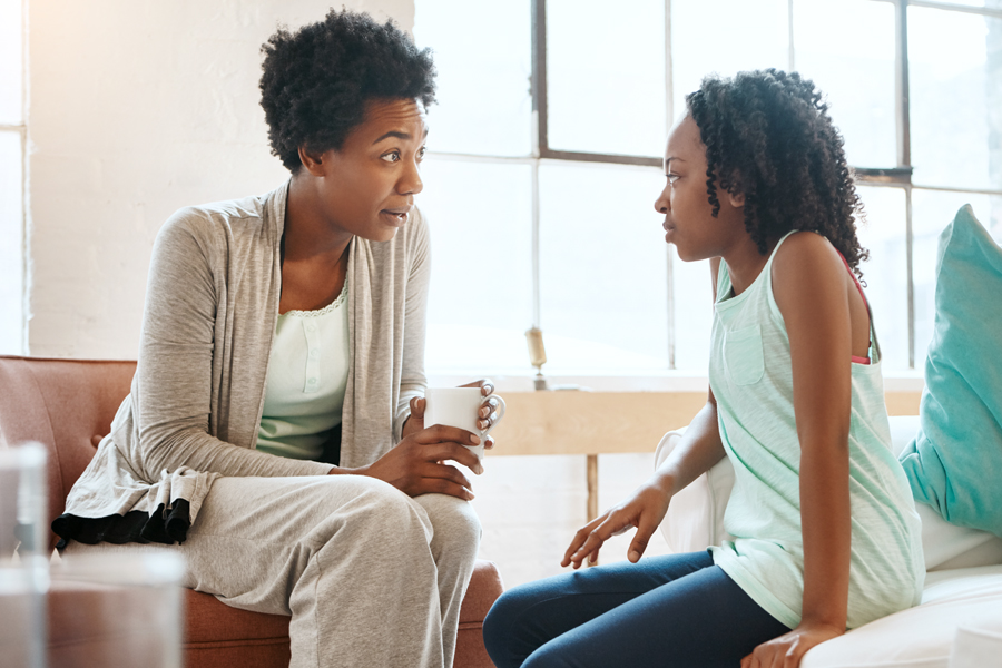 An adult woman talks with a young girl in a living room, showing attention and empathy as they share a serious but caring conversation.