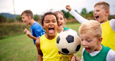 Group of young children run together on a soccer field with ball in the air, cheering