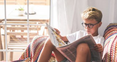 Beautiful young boy reading a newspaper at home. Blonde child reads news on a journal. Generation z child looking last-minute news on a paper magazine. information interest culture educational concept