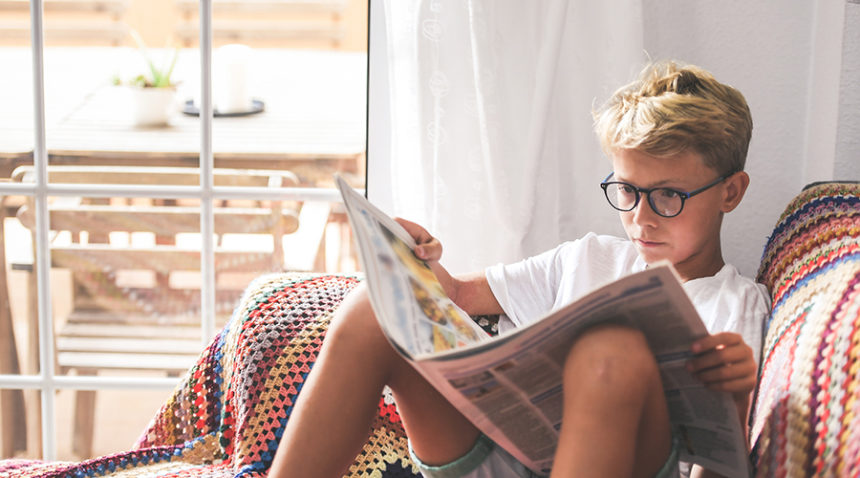 Beautiful young boy reading a newspaper at home. Blonde child reads news on a journal. Generation z child looking last-minute news on a paper magazine. information interest culture educational concept