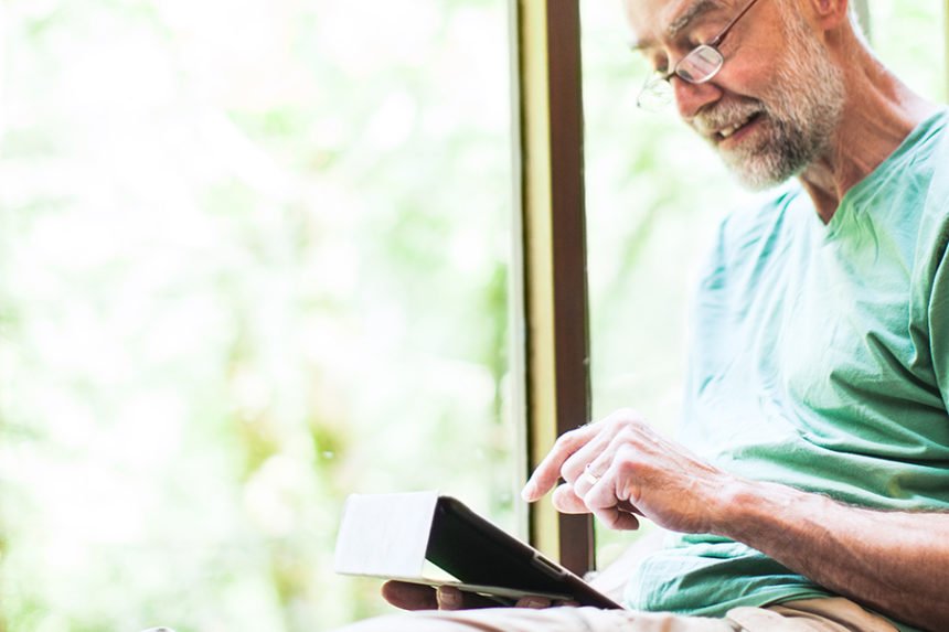 older man sitting by window, looking at tablet and smiling