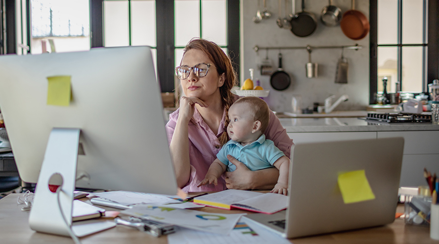 mom sitting at messy kitchen counter, looking at computer screen, holding infant on her lap