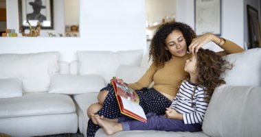 mom sitting on couch with young daugher, playing with her hair while she reads her a story