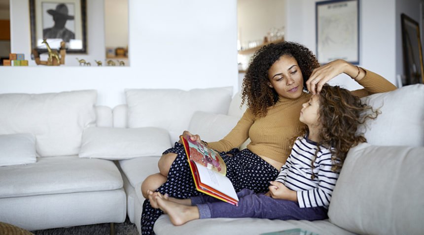 mom sitting on couch with young daugher, playing with her hair while she reads her a story