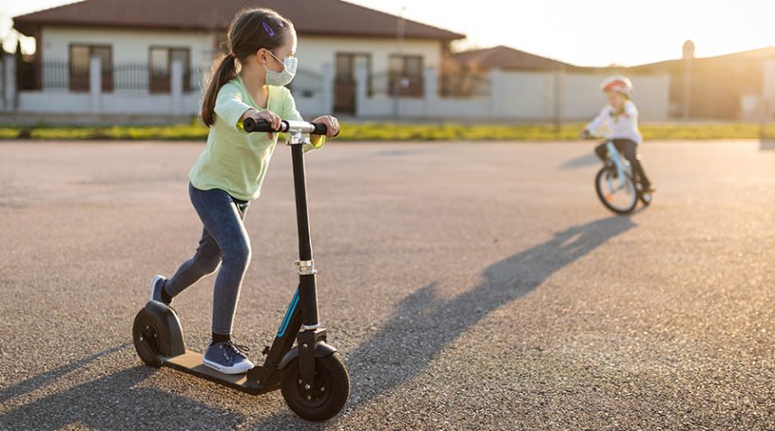 Two children play in cul-de-sac; girl wearing face mask rides scooter while a boy without mask rides bike