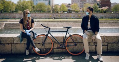 young woman and young man, both wearing face masks and gloves, sit on wall, chatting, with bicycle in between them