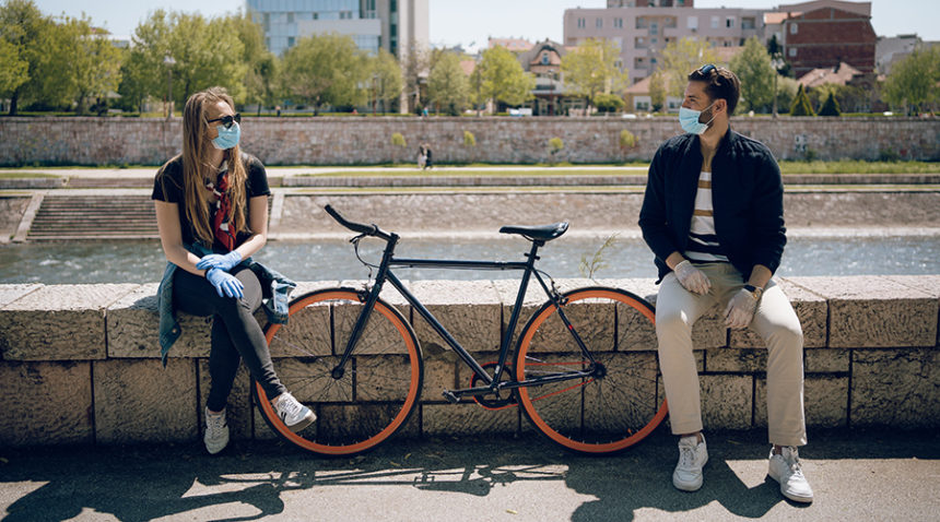 young woman and young man, both wearing face masks and gloves, sit on wall, chatting, with bicycle in between them