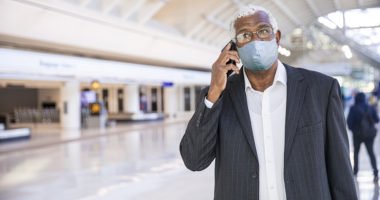 Businessman in airport lobby, talks on cellphone while wearing face mask