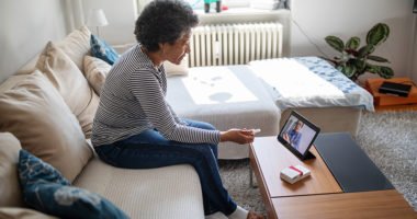 older woman sitting on couch in living room, video conferencing with a provider.