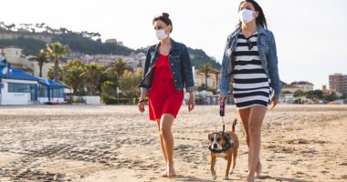 two women walk on beach, wearing face masks, as one walks dog