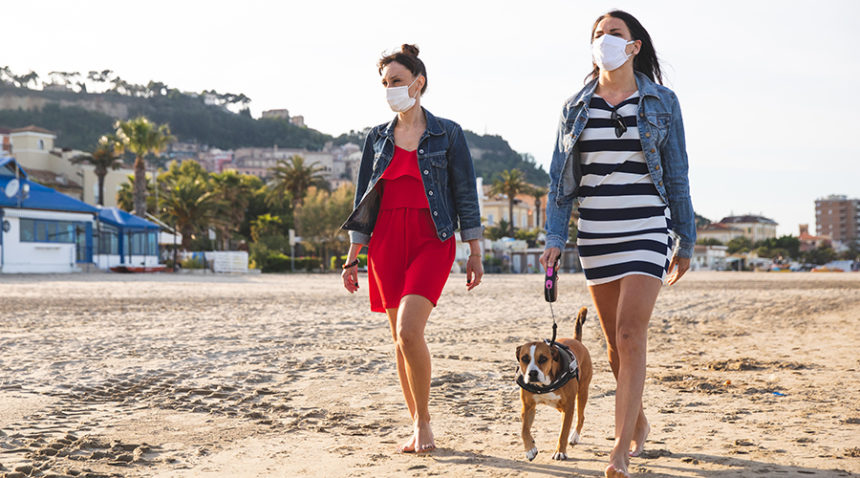 two women walk on beach, wearing face masks, as one walks dog