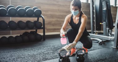 woman wearing fitness clothes, mask and gloves, sanitizes a kettleball weight in a gym