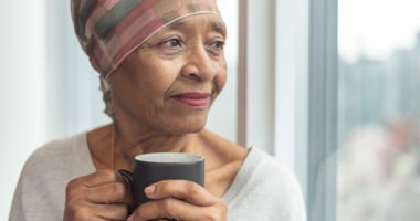 older woman wearing head wrap, holding coffee mug, looks out of window