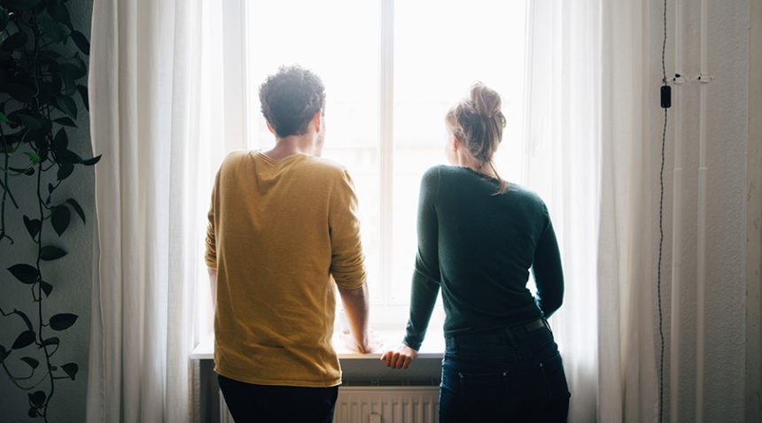 Two adults in a dark room look out of a window, backs to the camera