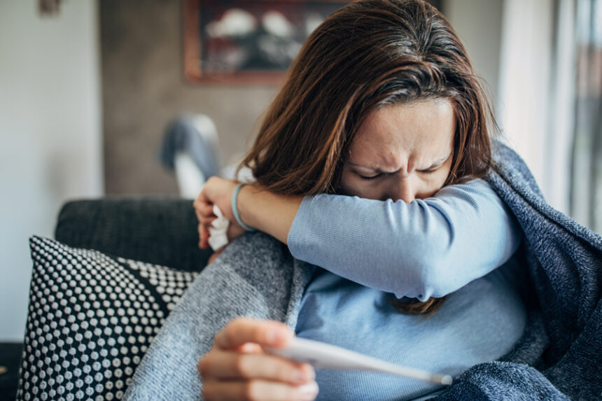 Woman sneezing into her arm while holding a thermometer