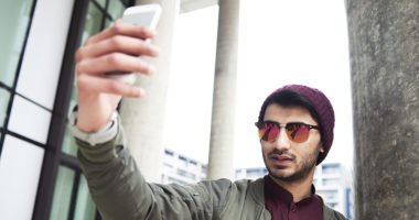 young man wearing sunglasses and beanie hat holds phone up to take selfie
