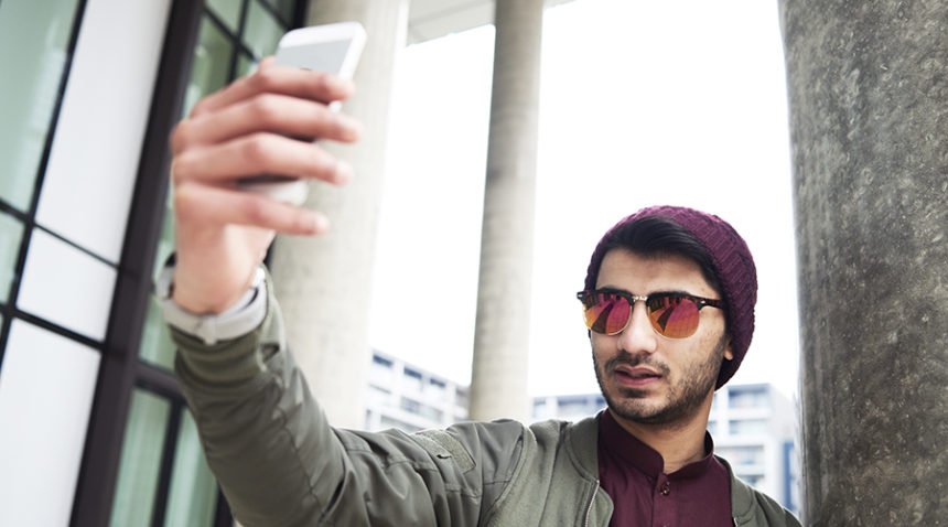 young man wearing sunglasses and beanie hat holds phone up to take selfie