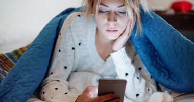 young woman lays under blanket, looking at smartphone