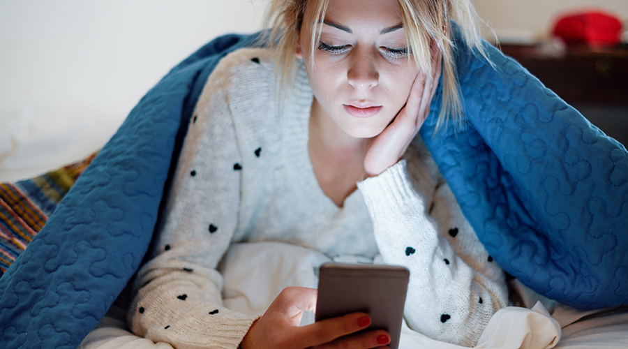 young woman lays under blanket, looking at smartphone