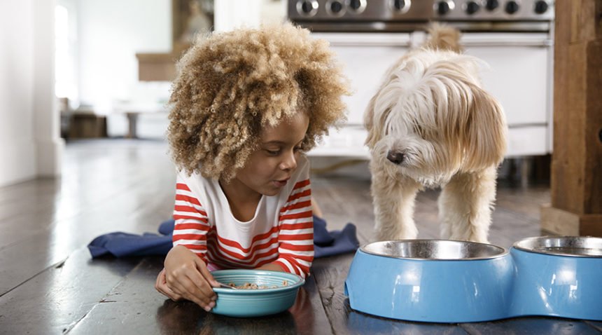 A little girl lays on her belly next to her dog, both in front of the dog's food bowl