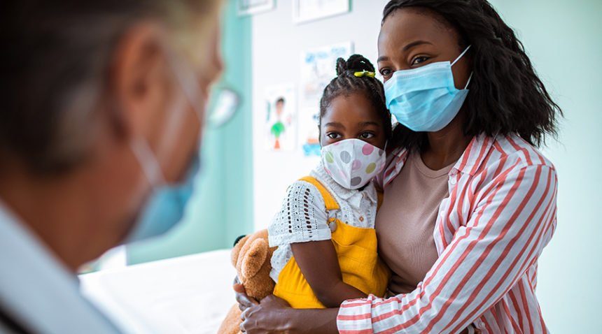 Mom and young daughter, wearing masks, talk to their pediatrician