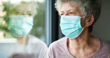 older woman wears a face mask while she looks out the window