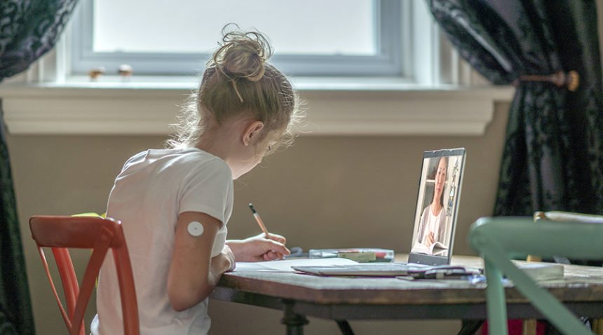 girl wearing a diabetes pump in her arm takes notes while watching a virtual class at her dining room table.
