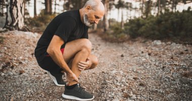 Senior man holding knee by hands and suffering with pain, kneeling on outdoor running trail.