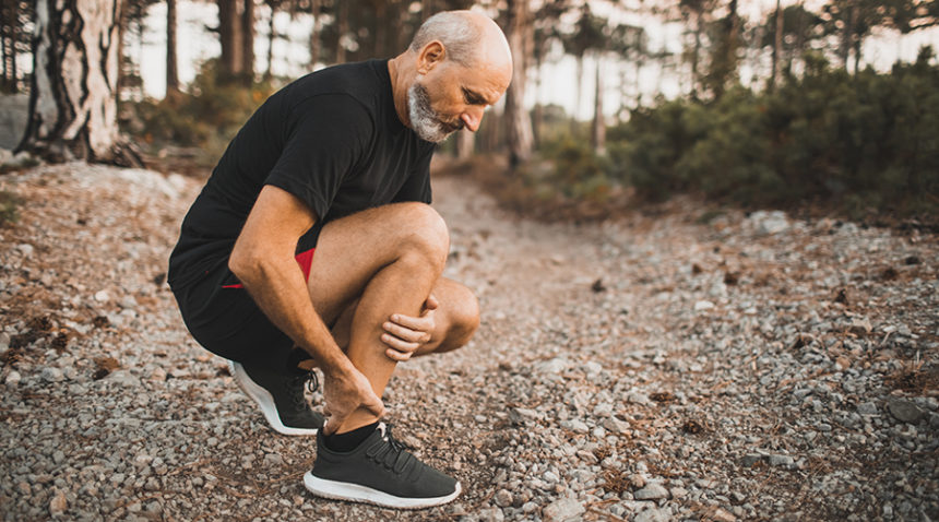 Senior man holding knee by hands and suffering with pain, kneeling on outdoor running trail.