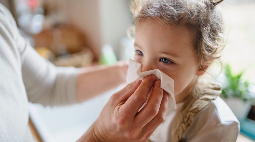 parent holds tissue, blowing nose of small sick daughter indoors at home.