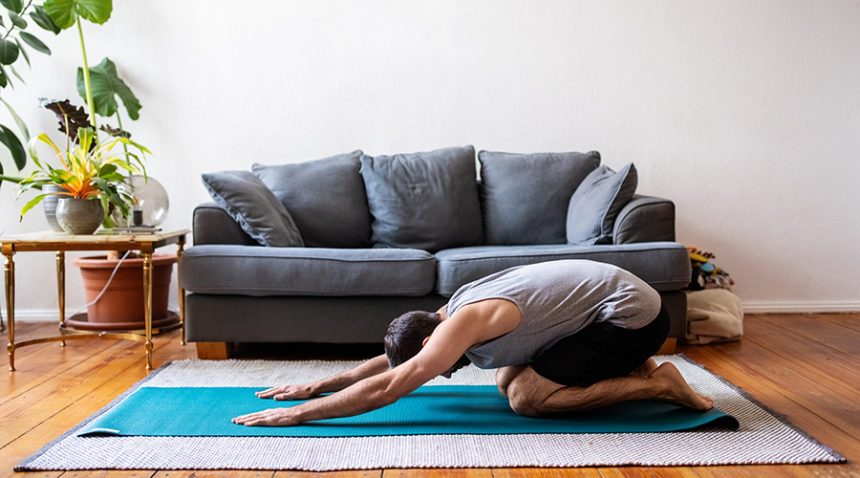 Side view of a mid adult male working out at home. Man doing childs pose yoga on exercise mat indoors