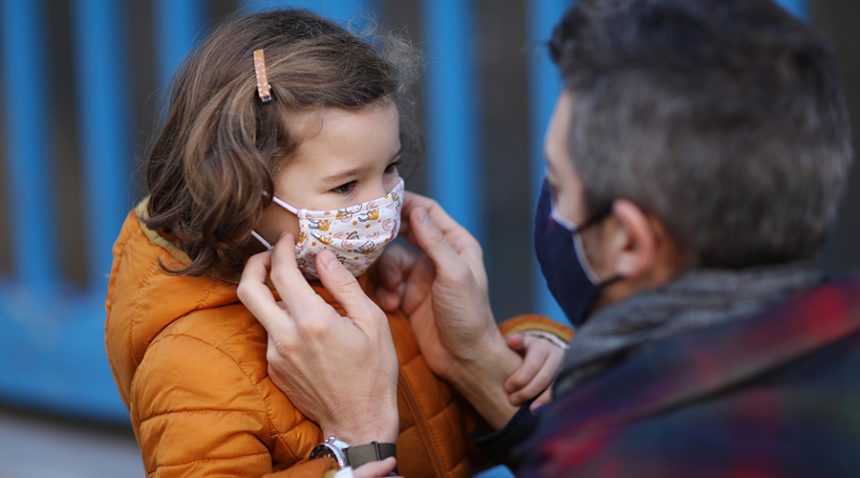 A father putting a protective face mask on his little daughter in front of school