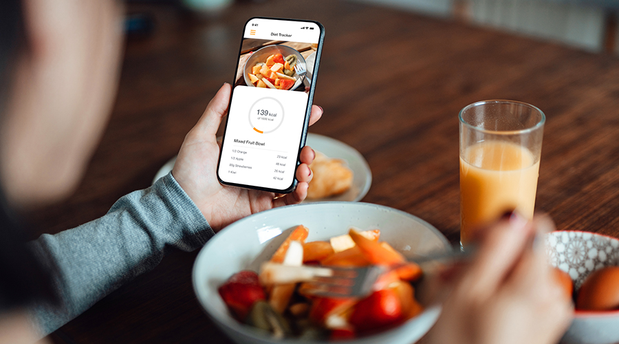 Over the shoulder shot of young woman using mobile app to track nutrition and count calories with smartphone while eating breakfast.