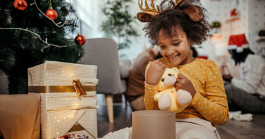 little girl opens present of stuffed animal next to Christmas tree