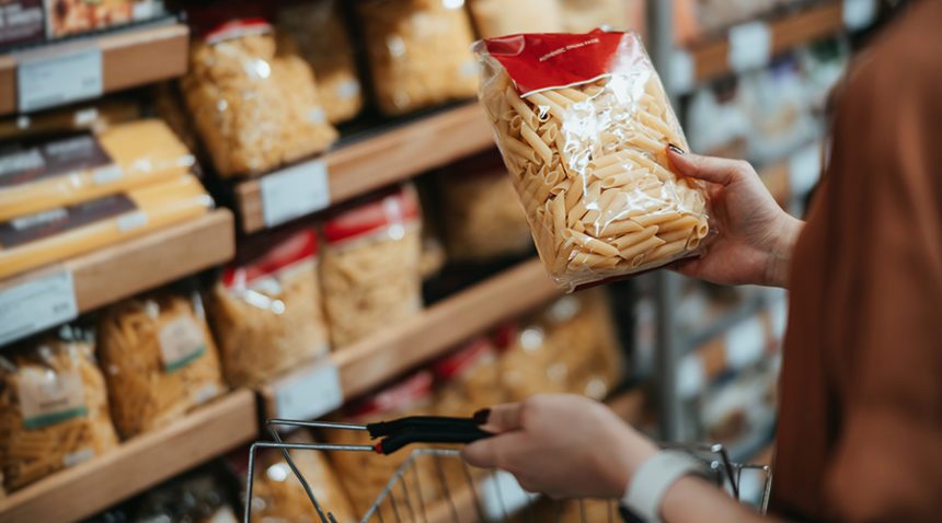 woman carrying a shopping basket, grocery shopping in supermarket, close up of her hand choosing a pack of organic pasta along the aisle.