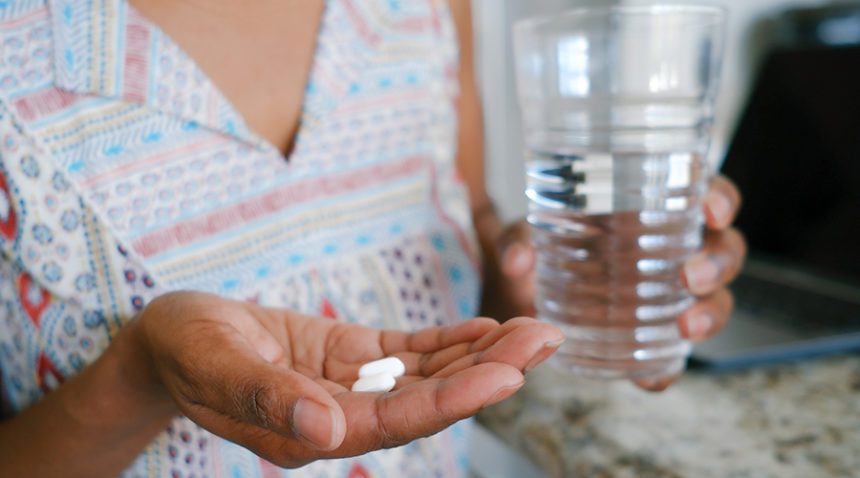 Close-up of unrecognizable woman in kitchen holding medication in one hand and a glass of water in the other.