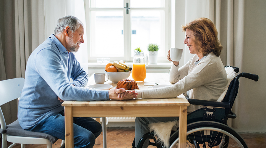 A woman in a wheelchair has breakfast or lunch with her husband, holding hands.