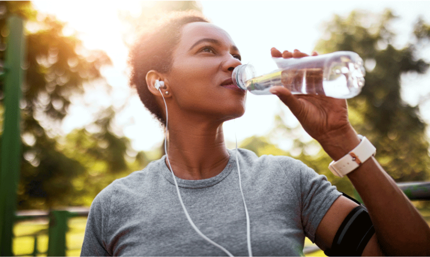 20s-30s-something woman takes a drink of water while listening to headphones outside, implied she is out for a walk or run